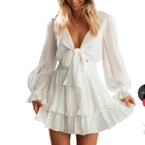 Vit klänning med knyt och volang - Säljer en superfin vit klänning med knyt framtill och volangdetaljer. Den har långa, genomskinliga ärmar och en kort längd. Klänningen är i ett lätt och luftigt material, vilket gör den idealisk för varma dagar. Perfekt till STUDENT!!👩🏼‍🎓💕Aldrig använd bara testat den på! Köpt för original priset.