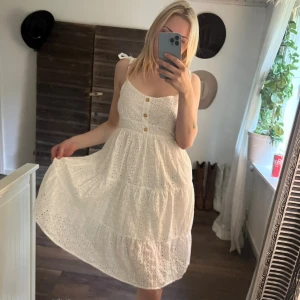 Vit somrig klänning med broderi - Superfin vit klänning med broderade hålmönster, knappar framtill och smala axelband som knyts. Klänningen har en lätt utsvängd kjol och dragkedja i sidan. Perfekt för varma dagar och har en romantisk vibe. Finns en lite skavank på bild tre. Men påverkar inte dragkedjan! 