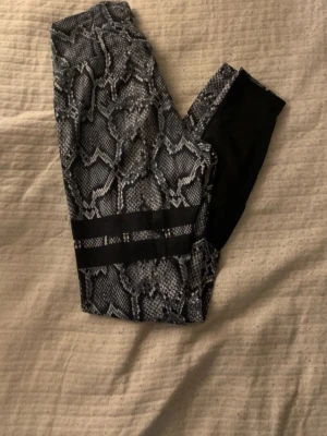STRONGER träningsleggings - Snygga träningsleggings från Stronger med svart och grått ormmönster. De är tillverkade i stretchigt syntetmaterial som passar perfekt för träning. Loggan är tryckt på sidan och designen är riktigt trendig.