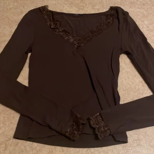 Brun långärmad topp med spetsdetaljer - Mörkbrun långärmad topp med v-ringning och dekorativa spetsdetaljer både vid halsringningen och vid ärmsluten. Tunn och mjuk i materialet, perfekt för dig som gillar en feminin och stilren look.