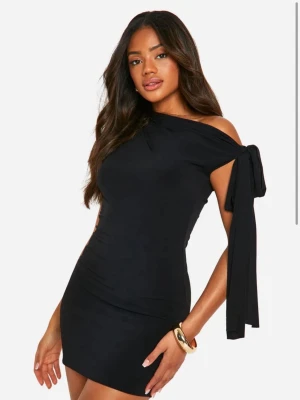 Svart one shoulder kortklänning - Snygg svart kortklänning med one shoulder-design och knyt-detalj på ärmen. Klänningen har aldrig använts och kommer med prislappen på. 