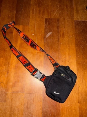 Svart axelväska från Nike - Säljer en svart axelväska från Nike med justerbart band i svart och orange med stora Nike-loggor. Väskan har dragkedja upptill och en mindre ficka framtill. Perfekt för att bära det viktigaste när du är på språng.