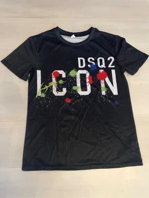 Svart DSQ2 ICON t-shirt med färgstänk - Svart t-shirt från DSQ2 med stor vit 'ICON'-text och färgstänk i rött, grönt och blått framtill. Klassisk rund hals och korta ärmar. Materialet känns lätt och syntetiskt, perfekt för en cool streetwear-look.
