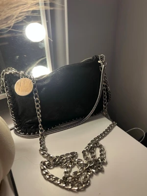 Svart axelväska kedja - Säljer en svart axelväska med grov silverkedja och dragkedja längs kanten. Väskan har en rund metallbricka som detalj och är tillverkad i skinnliknande material. Perfekt för dig som gillar en edgy stil och vill ha något som sticker ut.