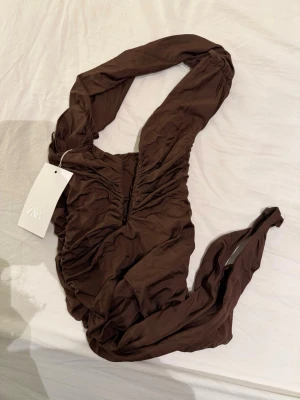 Brun omlott body från Zara - Säljer en brun omlott body från Zara i storlek M. Body  har en snygg omlottdesign med rynkade detaljer framtill och breda axelband. Materialet är mjukt och stretchigt, vilket gör den riktigt bekväm och smickrande.