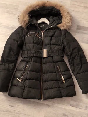 Svart pufferjacka från Hollies, strl 36 - Säljer en svart pufferjacka från Hollies med snyggt bälte i midjan och guldfärgade dragkedjor. Jackan har huva med fejkpäls och leopardmönstrat foder. Perfekt för kalla dagar och riktigt skön passform. Flera fickor med dragkedja och quiltad design.