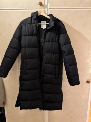 Svart lång pufferjacka från Kappahl - Svart pufferjacka från Kappahl i storlek 170. Jackan är lång, har huva, dragkedja framtill och slits på sidan med knappar i sidorna. säljer den då jag köpt ny vinterjacka och behöver rensa garderoben😅