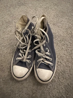 Mörkblå Converse All Star high tops - Klassiska mörkblå Converse Chuck Taylor All Star high tops med vita gummisulor och tåhättor. Skorna har vita snören, silverfärgade öljetter och den ikoniska loggan på sidan. Tillverkade i textil med vit kontrastsöm och rund tå.