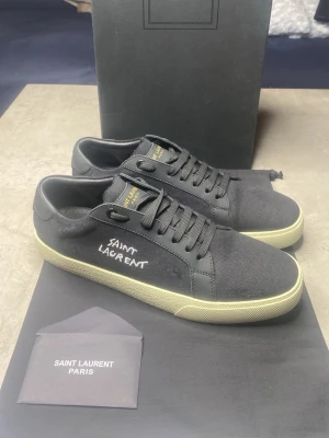 Svarta Saint Laurent sneakers - Snygga svarta sneakers från Saint Laurent med handskriven logga på sidan. Skorna har ovandel i canvas och detaljer i skinn, samt en vit platt gummisula. Klassisk låg modell med snörning och diskreta Saint Laurent-detaljer på plös och sula.