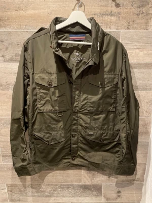 Snoot jacka  - Säljer en olivgrön vindjacka från Snoot med flera stora fickor framtill, axelklaffar och snygga detaljer som dragkedja vid kragen. Jackan är tillverkad i polyester och har en klassisk militärinspirerad look. Perfekt för dig som gillar stilren och funktionell design.