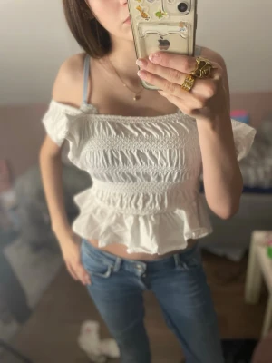 Vit offshoulder blus från Zara - Supersöt vit blus från Zara med offshoulder och volangkant nertill. Smockad överdel som ger en tight passform och kort, croppad modell. Perfekt för sommarens alla tillfällen och lätt att matcha med jeans eller kjol.