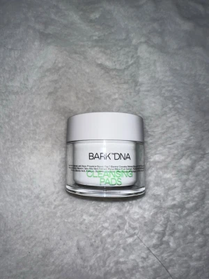 BARK DNA Cleansing Pads - Runda rengöringspads från BARK DNA i en transparent burk med vit lock. Padsen är designade för att effektivt ta bort smink och rengöra huden. Förpackningen är stilren och modern med svart och grön text på framsidan.
