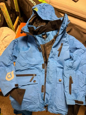 Blå skidjacka från Kappahl   WOXO - Blå skidjacka  från Kappahl WOXO 720 med flera fickor, huva och svarta detaljer. Snölås och Liftkortsficka på armen. Perfekt för dig som gillar funktionella och snygga ytterplagg. Snygg logga på ärmen och justerbara muddar.