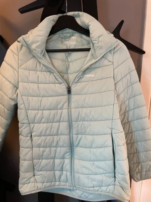 Ljusblå pufferjacka från Everest - Snygg ljusblå pufferjacka från Everest med quiltad design och dragkedja framtill. Jackan har hög krage och två sidofickor med dragkedja. Perfekt för kyliga dagar och enkel att matcha med olika outfits.