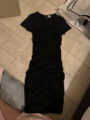 Svart ribbad klänning från Divided - Svart ribbad klänning från Divided med kort ärm och snygg rynkdetalj längs hela framsidan. Modellen är kroppsnära och har en rund halsringning. Perfekt för dig som gillar en enkel men trendig look. Finns ingen prislapp men har aldrig använt eller tvättat den