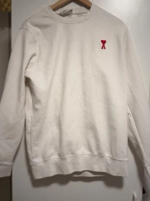 Vit sweatshirt från Ami Alexandre Mattiussi - Vit sweatshirt från Ami Alexandre Mattiussi med rött broderat hjärta och bokstaven A på bröstet samt röd AMI-text på ryggen. Rund hals och långa ärmar. Tillverkad i mjuk bomull för skön komfort och clean look.