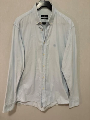 Ljusblå skjorta från Henri Lloyd - Klassisk ljusblå skjorta från Henri Lloyd med button-down krage och diskret logga broderad på bröstet. Skjortan har långa ärmar, normal passform och stängs med vita knappar framtill. Perfekt till jeans eller under kavaj.