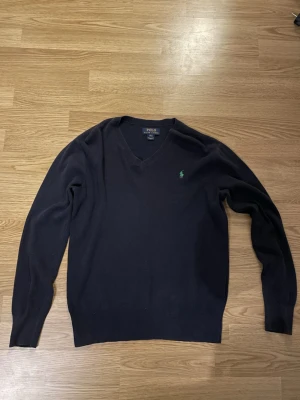 Mörkblå stickad tröja Polo Ralph Lauren - Snygg mörkblå stickad tröja från Polo Ralph Lauren med v-ringad hals och klassisk grön logga broderad på bröstet. Tröjan har långärmad passform och ribbade muddar vid ärmslut och nederkant. Perfekt för dig som gillar stilrena och tidlösa plagg. Barnstorlek L så passar S möjligen XS