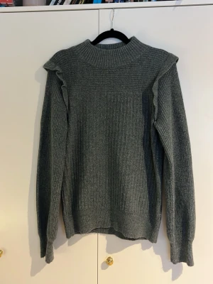 Stickad tröja med volangdetalj - Mysig grå/grön stickad tröja med ribbad struktur och volangdetaljer längs axlarna. Tröjan har hög hals och långa ärmar med breda muddar. Perfekt för kyliga dagar och enkel att styla till olika outfits.