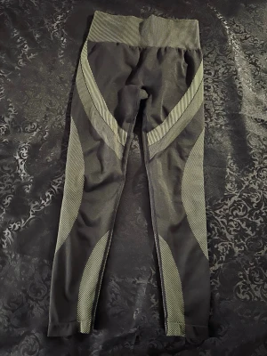 Svarta leggings med mönstrade detaljer - Säljer ett par svarta leggings med ljusgröna mönstrade partier längs benen och bred ribbad midja. Tillverkade i stretchigt material (90% polyamid, 10% elastan) som sitter tight och bekvämt. Perfekta för träning eller chill dagar.
