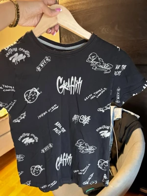 Svart graffiti t-shirt från Max - Cool svart t-shirt från Max med vit graffiti-inspirerad print över hela plagget. T-shirten har rund hals och korta ärmar. Materialet är mjuk bomull som är skönt mot huden. Perfekt för dig som gillar streetstyle och vill sticka ut med ett unikt tryck.