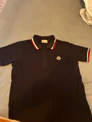 Svart Moncler piké med röd-vit krage - Säljer en svart pikétröja från Moncler med klassisk krage i rött och vitt, samt broderad Moncler-logga på bröstet. Tröjan har korta ärmar och knäppning med tre svarta knappar framtill. Kragen har Moncler-text broderad i vitt.