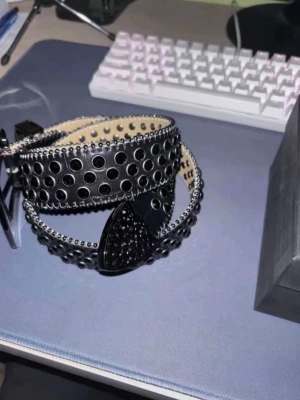 Svart b.b. simon skärp med nitar - Coolt svart b.b. simon skärp med glittriga nitar och stora metallhål längs hela bandet. Spännet är dekorerat med svarta stenar och har en unik form. Skärpet har en croco-textur och detaljerade kanter som ger extra edge till din outfit.