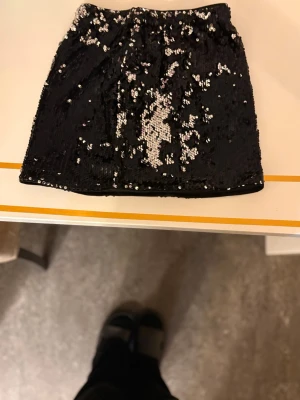 Svart paljettkjol med silverdetaljer - Säljer en kort svart kjol täckt med glittriga paljetter i svart och silver. Kjolen har rak passform och resår i midjan för en bekväm känsla. Perfekt för dig som vill sticka ut med en glittrig look. Använd 1 gång💕