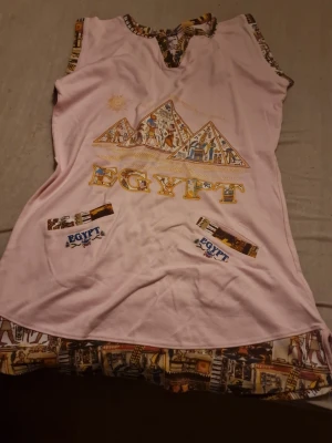 Rosa Egypten-klänning från Maria - Unik rosa kortklänning från Maria i 100% bomull med färgglada egyptiska motiv och texten 'EGYPT' framtill. Klänningen har ärmlös design, dekorativa kanter och tryckta detaljer med pyramider och hieroglyfer. Perfekt för dig som gillar statement-plagg och coola prints. En traditionell egyptisk klänning som kallas "galabiya", perfekt för en resa till Egypten för att visa respekt för kultur eller om du själv är egyptier/nordafrikan. Priset som står gäller, kan möjligtvis diskuteras🫶