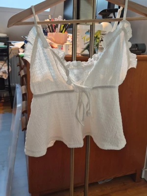 Vit linnetopp med volang och knyt - Säljer en söt vit linnetopp med tunna axelband och volangdetaljer längs halsringningen. Toppen har en knytning framtill och är tillverkad i ett lätt, krusat material som ger en luftig känsla. Perfekt för varma dagar och sommarhäng.