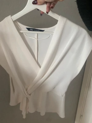 Vit omlottblus från Zara - Säljer en stilren vit omlottblus från Zara med knyt i sidan och v-ringad hals. Blusen är ärmlös och har en mjuk, lätt struktur som ger en snygg siluett. Perfekt att matcha med jeans eller kjol för en fräsch look. Använd max 2 gånger, nyskick.