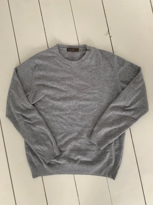 Grå ulltröja från Oscar Jacobson - Säljer en stilren grå ulltröja från Oscar Jacobson, tillverkad i Italien. Tröjan har rund hals, ribbade muddar och är långärmad. Perfekt för dig som gillar klassisk och mjuk design med premiumkänsla.