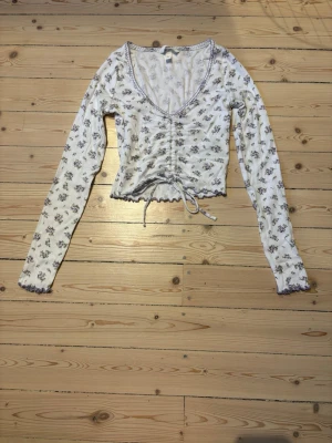 Blommig croppad långärmad topp - Säljer en vit croppad topp med lila blommönster och lång ärm. Toppen har v-ringning, snörning framtill och volangkant vid ärmslut och nederkant. Materialet känns mjukt och stretchigt, perfekt för dig som gillar romantiska detaljer och en feminin stil.