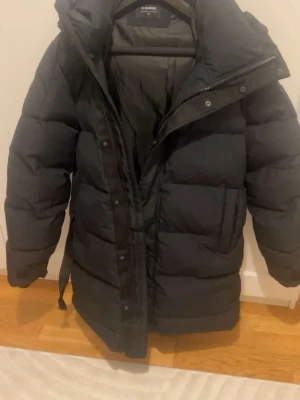 Svart pufferjacka från Everest XS - Svart pufferjacka från Everest i storlek XS, men den är ganska stor. Jackan har en hög krage, luva och dragkedja med knappar framtill. Den är quiltad och har en loose passform, perfekt för kalla dagar. Materialet är polyester och jackan är riktigt varm och skön. Köpte för 1999kr