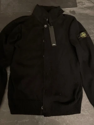 Stone island kofta  - Svart stickad kofta från Stone Island med ikonisk patch på ärmen. Koftan har huva, knappar framtill och dragkedja under knappslån. Perfekt för dig som gillar streetwear och vill ha något stilrent och bekvämt. Priset är inte hugget i sten:)