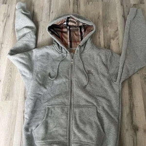 Grå Burberry hoodie med dragkedja - Snygg grå hoodie från Burberry. Tröjan har dragkedja, snörning i huvan och två stora fickor, Perfekt för dig som gillar sköna och exklusiva plagg. 