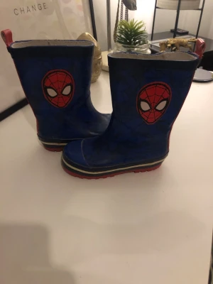 Blå Spiderman stövlar för barn st 24 - Säljer ett par blå stövlar med Spiderman-tryck på sidorna och röd text bak. Snygga röda sulor och detaljer i vitt och svart. Tillverkade i syntetmaterial, perfekta för regniga dagar. Cool design för alla Spiderman-fans!. St 24.