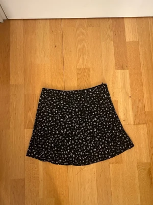 Svart blommig kjol från H&M Divided - Supersöt svart kortkjol från H&M Divided med små blommor i grön och rosa. Kjolen har en lätt utställd form och är gjord i ett mjukt, tunt material som känns skönt mot huden. Perfekt att styla med en enkel topp eller stickad tröja.