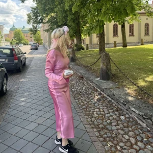 Rosa långklänning i satin med öppen rygg och rosett - Säljer eftertraktade klänningen i satin från gina som ej går att köpa längre. Har använt den endast 1 gång på midsommar samma klänning som saga stq har i fint skick kontakta gärna för fler bilder🩷🫶🏼 har en liten repa bak i ryggen men inget hål