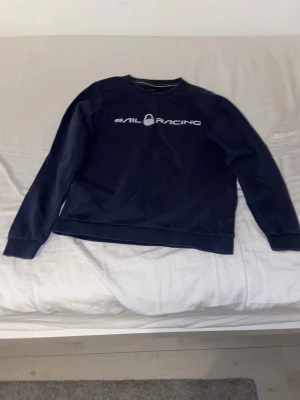 Mörkblå Sail Racing sweatshirt - Snygg mörkblå sweatshirt från Sail Racing med vit logga tryckt på bröstet och en mindre logga i nacken. Tröjan har rund hals, långa ärmar och ribbade muddar. Perfekt för dig som gillar stilren design och sportiga märken.
