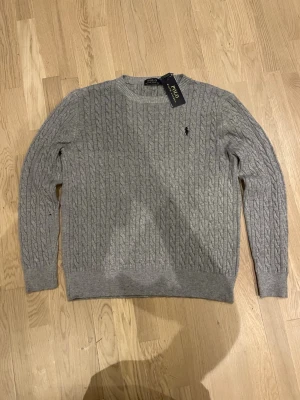 Grå kabelstickad tröja Polo Ralph Lauren - Snygg grå kabelstickad tröja från Polo Ralph Lauren med klassisk rund hals och diskret logga på bröstet. Tröjan har långärmade ribbade muddar och är tillverkad i mjukt material som passar perfekt för kyliga dagar.