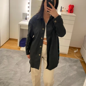 Svart kappa i skinn imitation från NA-KD med bälte - Svart rock från NA-KD i storlek 34. Jackan har klassisk krage, knappar framtill och ett snyggt bälte med metallring i midjan. Långärmad och rak passform, perfekt för dig som gillar stilrena och tidlösa plagg.