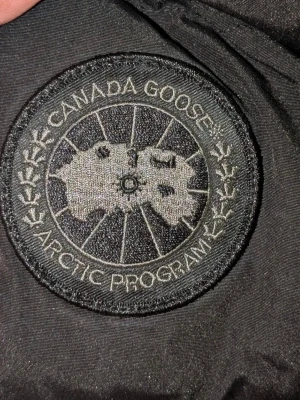 Svart pufferjacka från Canada Goose XL - Svart pufferjacka från Canada Goose i storlek XL. Jackan har huva, ribbade muddar och klassisk logotyp på ärmen. Fylld med dun och har dragkedja samt knappar framtill. Perfekt för kalla dagar och har en clean, stilren look.