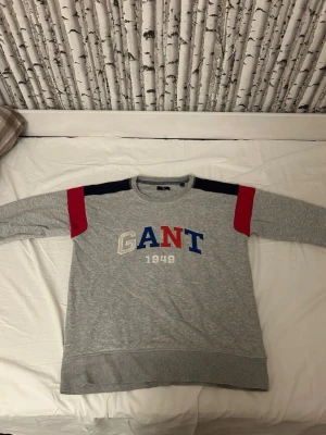 Grå sweatshirt från GANT med färgblock - Snygg grå sweatshirt från GANT med broderad logga och årtal på bröstet. Tröjan har färgblock i marinblått och rött på axlarna samt långa ärmar och ribbade muddar. Perfekt för dig som gillar klassisk streetstil och vill ha något bekvämt.