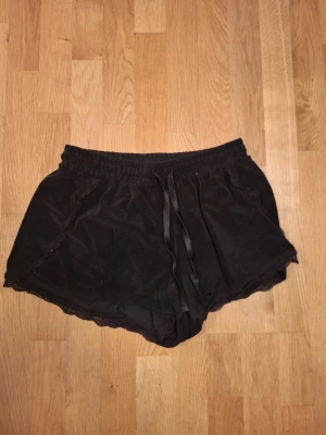 Svarta pyjamasshorts med spets från Isabella - Svarta shorts från Isabella i storlek M, gjorda av mjuk polyester och elastan. De har en loose passform, snörning i midjan och fina spetsdetaljer längs kanterna. Perfekta för chill dagar eller när du vill ha något bekvämt och snyggt. Passar även S.