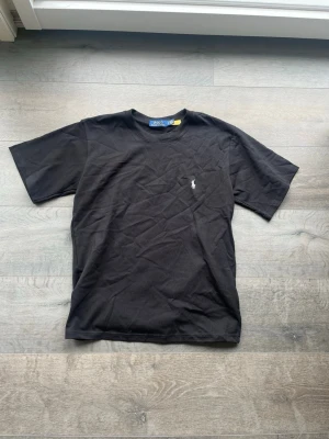 Svart t-shirt från Polo Ralph Lauren - Svart t-shirt från Polo Ralph Lauren med klassisk passform och korta ärmar. Ikoniska vita broderade loggan på bröstet. Tillverkad i mjuk bomull som känns skön mot huden. Perfekt till jeans eller shorts för en clean och stilren look.