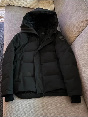 Svart pufferjacka från Canada Goose - Svart pufferjacka från Canada Goose med huva och klassisk patch på ärmen. Jackan har en stilren look med dragkedja och knappar framtill, samt ribbade muddar vid ärmslut. Perfekt för kalla dagar och har en riktigt schysst passform.Den är helt äkta och finns kvitto pris kan diskuteras vid snabb affär 