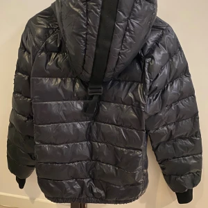 Svart pufferjacka Peak Performance - Säljer en svart pufferjacka från Peak Performance med huva och snygga detaljer som logga på ärmen och band med broderad text på ryggen. Jackan har dragkedja framtill och är tillverkad i glansig polyester. Perfekt för kalla dagar och har en schysst oversized look.