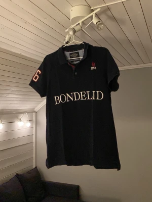 Svart Bondelid t-shirt med tryck - Säljer en svart t-shirt från Bondelid med stor vit logotyp på bröstet och röd detalj. T-shirten har korta ärmar, klassisk krage och knappslå. Perfekt för dig som gillar stilren design och tydliga detaljer.