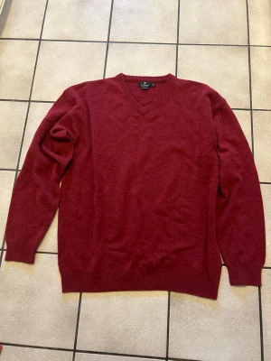 Vinröd v-ringad tröja MC Gordon XL 100% ull - Vinröd v-ringad tröja från MC Gordon i storlek XL, tillverkad av 100% lammull med Woolmark-märkning. Tröjan har långa ärmar och ribbade muddar vid hals, ärmslut och nederkant. Perfekt för dig som gillar klassisk stil och vill ha något mjukt och varmt.